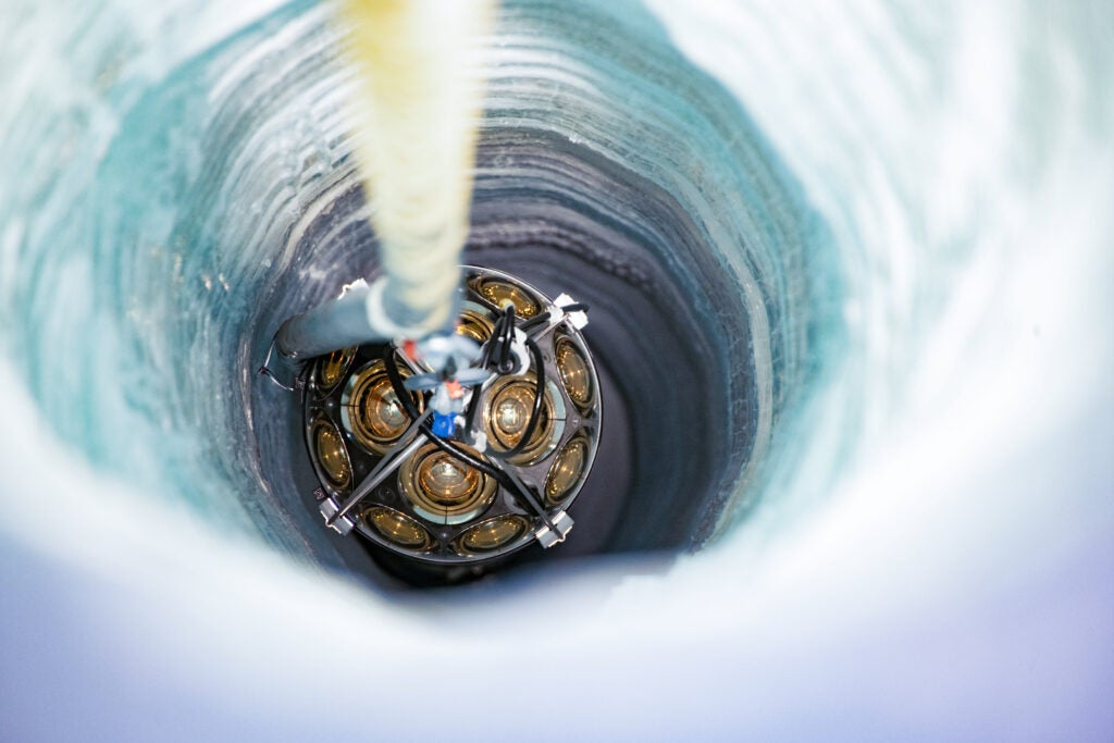 A device used to detect neutrinos is lowered into a hole where layers of blue and gray ice are visible in the hole walls.