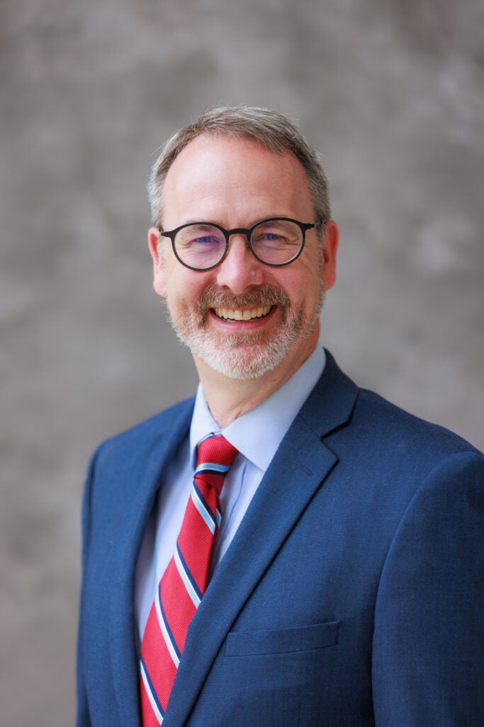 Gregory Thompson wearing a blue blazer and striped tie in front of a gray background. 