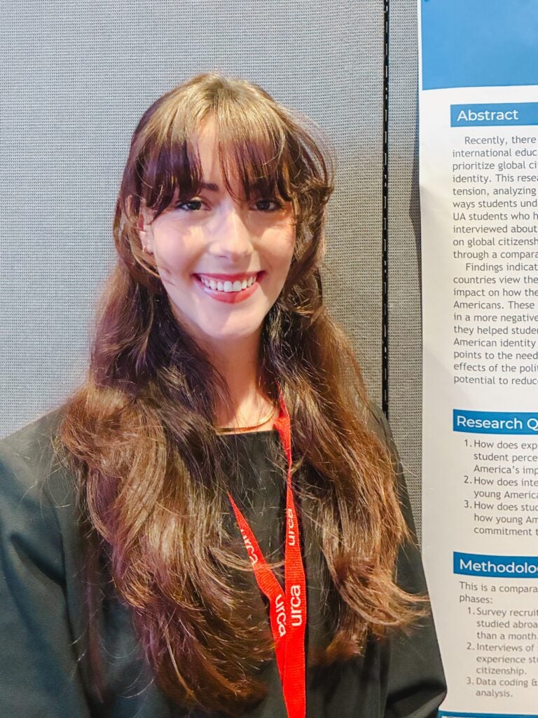 Young woman with light skin and very long brown hair, wearing a long sleeved black dress. She is standing by her research poster. 