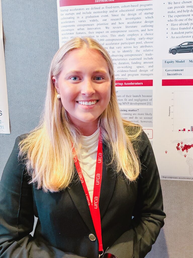 Young woman with light skin and long blonde hair, wearing a black blazer and white blouse, standing in front of her research poster.
