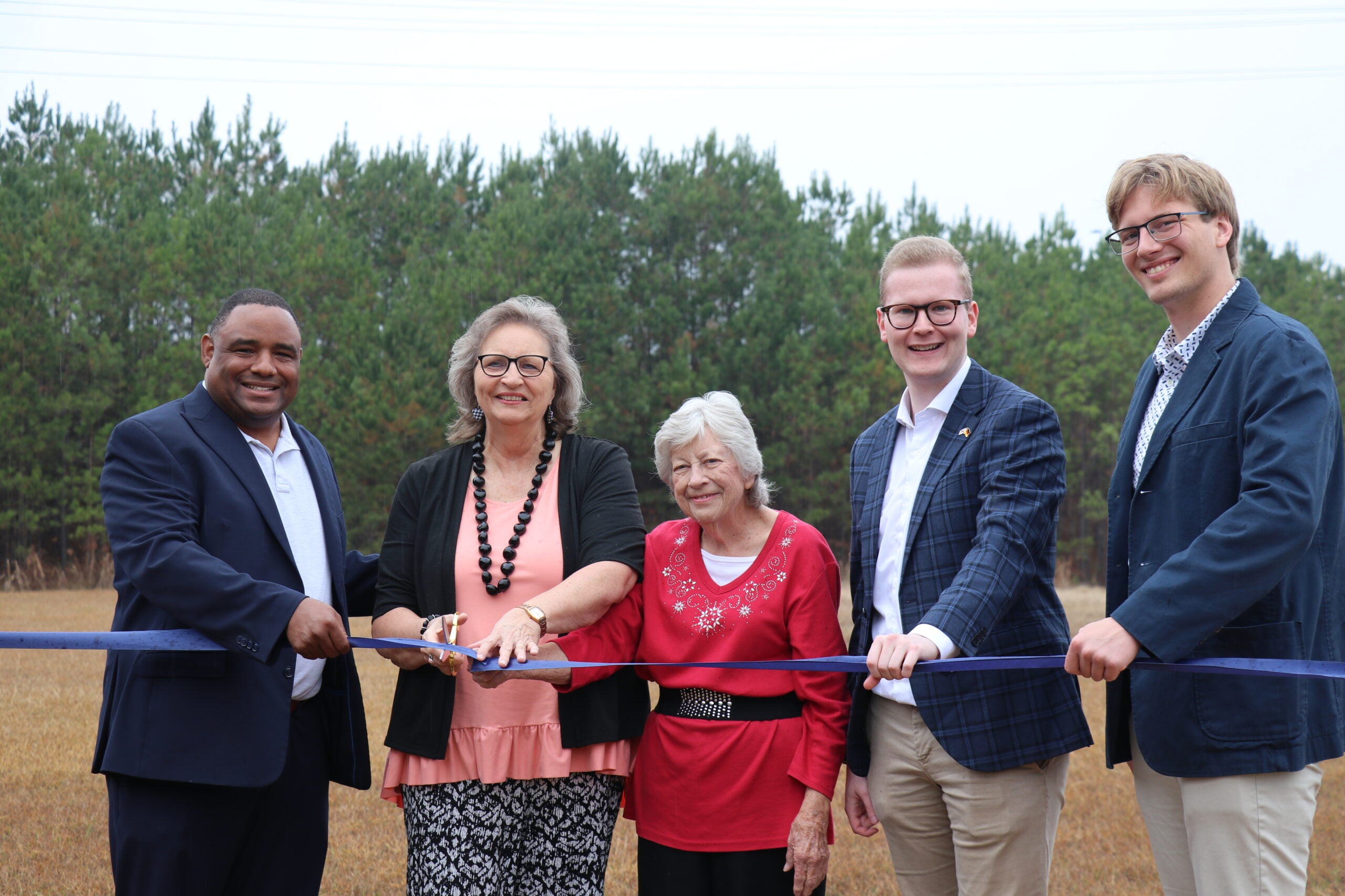 Five people stand behind a blue ribbon for a ribbon-cutting event. Vance mayor Brenda Morrison holds scissors, and the other participants are resting one hand on the ribbon. They are outdoors with pine trees in the background.