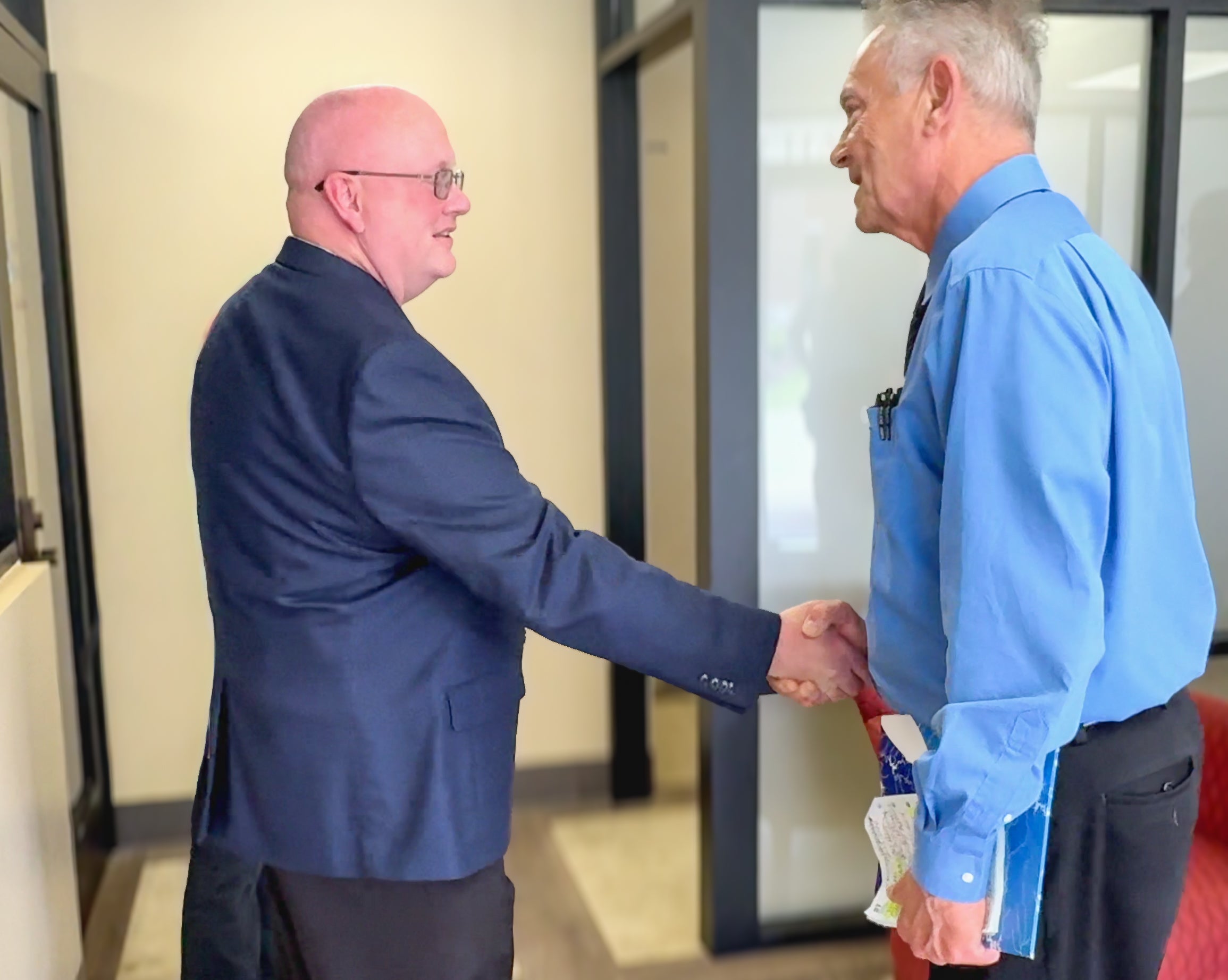 Two men shake hands in a hallway. The man on the left is bald and wears glasses and a navy blazer. The man on the right has gray hair and wears a blue dress shirt.