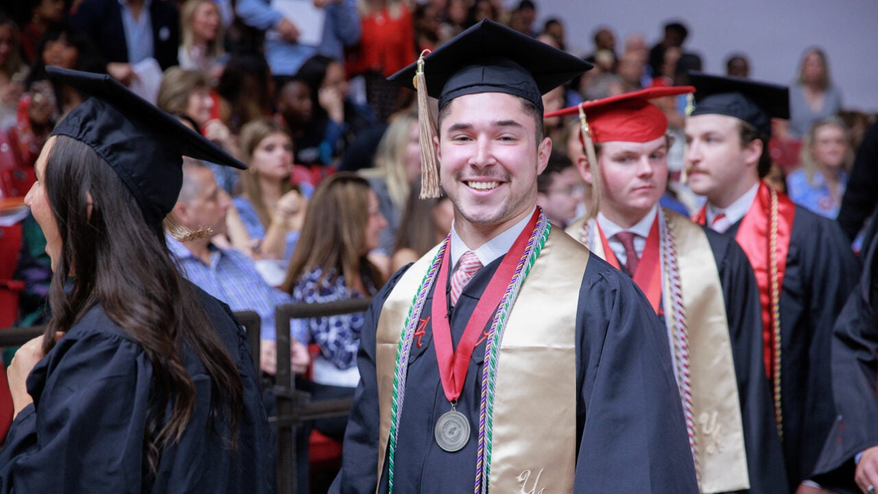 a young man smiles proudly for the camera while walking to his seat at commencement.