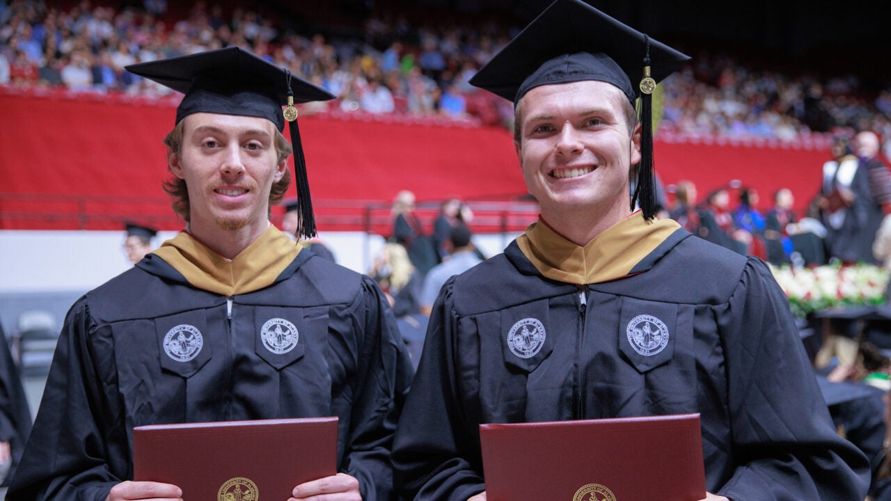 two young men smile for the camera while holding up their degrees