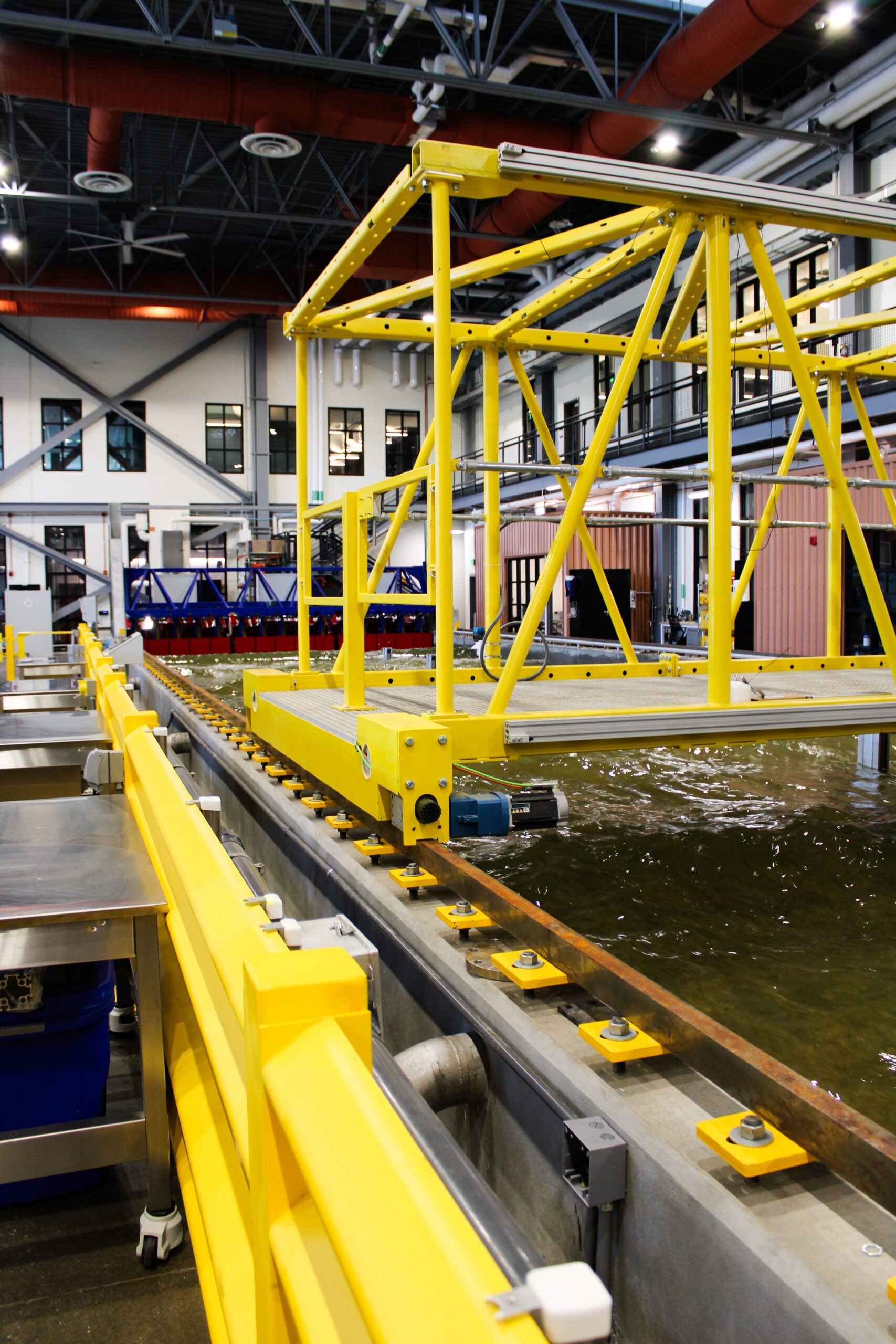 A large indoor hydraulic testing facility with an active water tank in operation. A blue metal bridge-like structure spans the width of the turbulent water. Yellow track hardware lines the foreground and small sensor instruments are visible in the water. A staircase, upper-level walkway and wood-paneled structure are visible in the background.