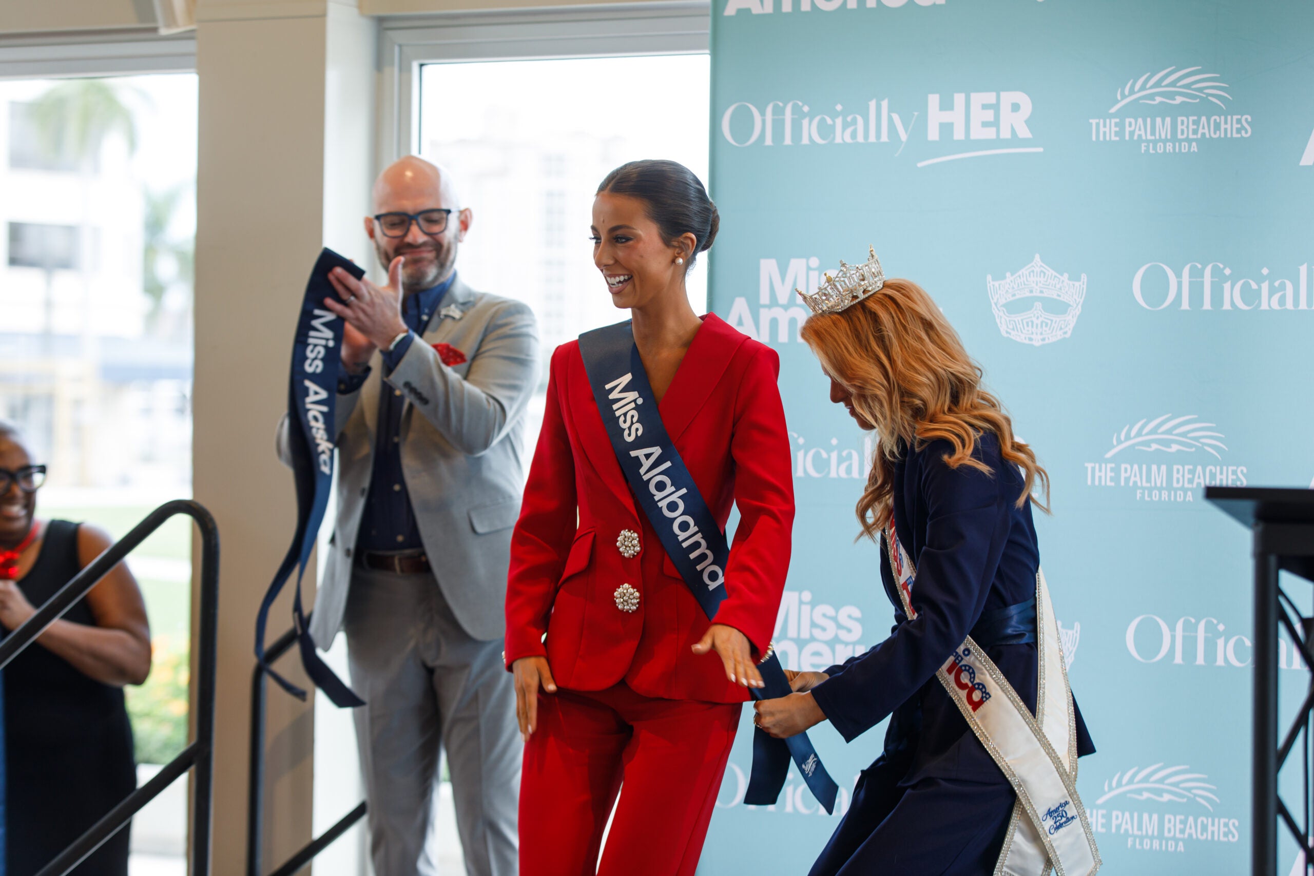 Tilghman smiling in a red pantsuit while Miss America drapes Miss Alabama sash over Tilghman's torso.