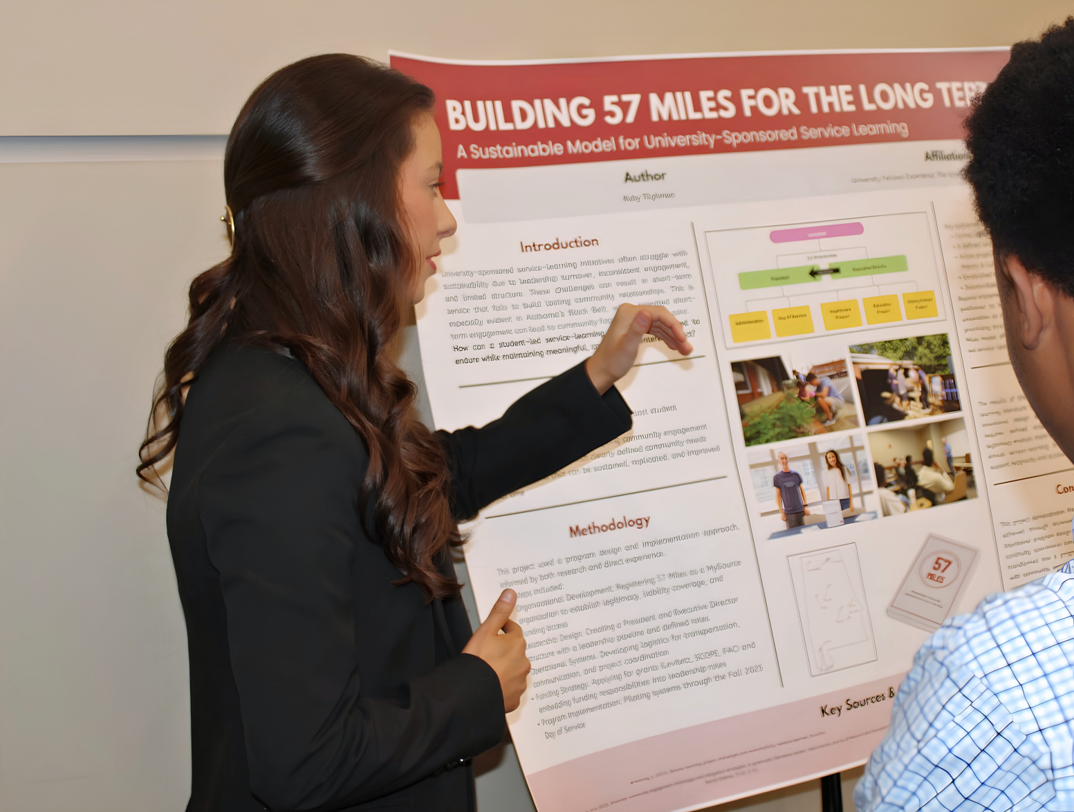 Tilghman in a black suit pointing to a red and white poster.