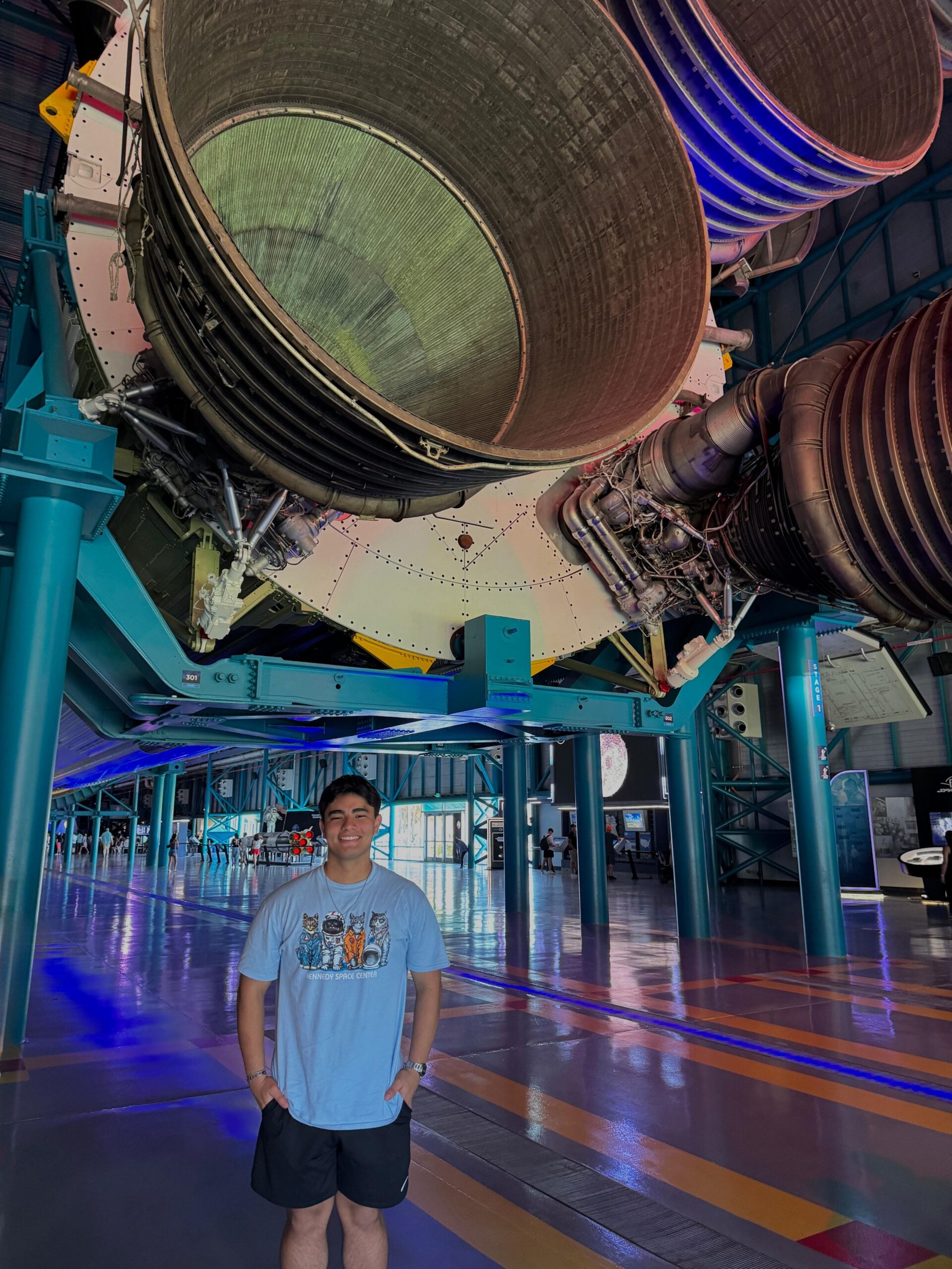 Fernando Salinas stands beneath the engines of a large rocket exhibit during his participation in the International Space Weather Camp, which included a visit to the Kennedy Space Center in Florida, shown here.